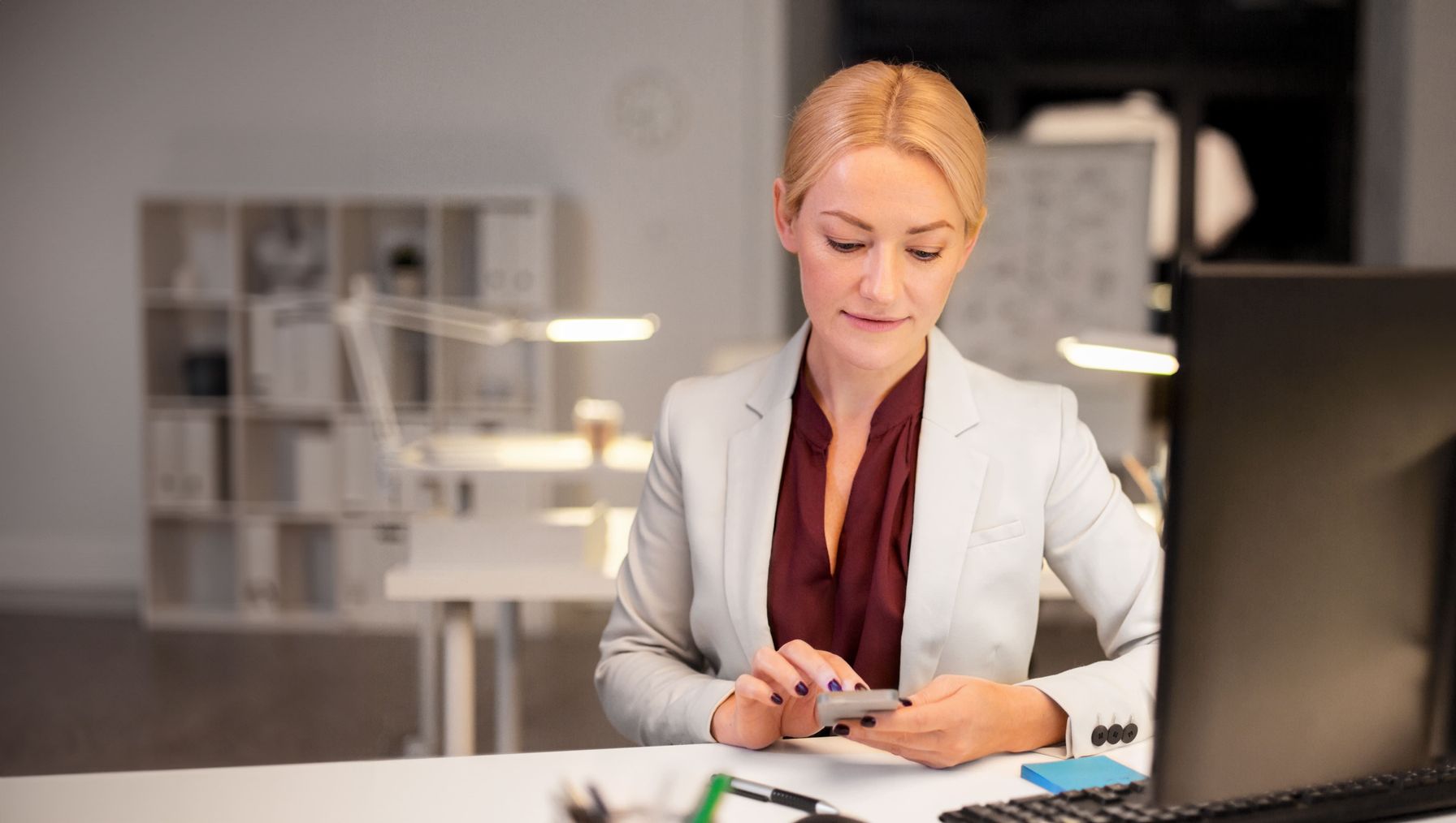 A professional woman in a blazer using her smartphone while working late at her desk in a modern office.