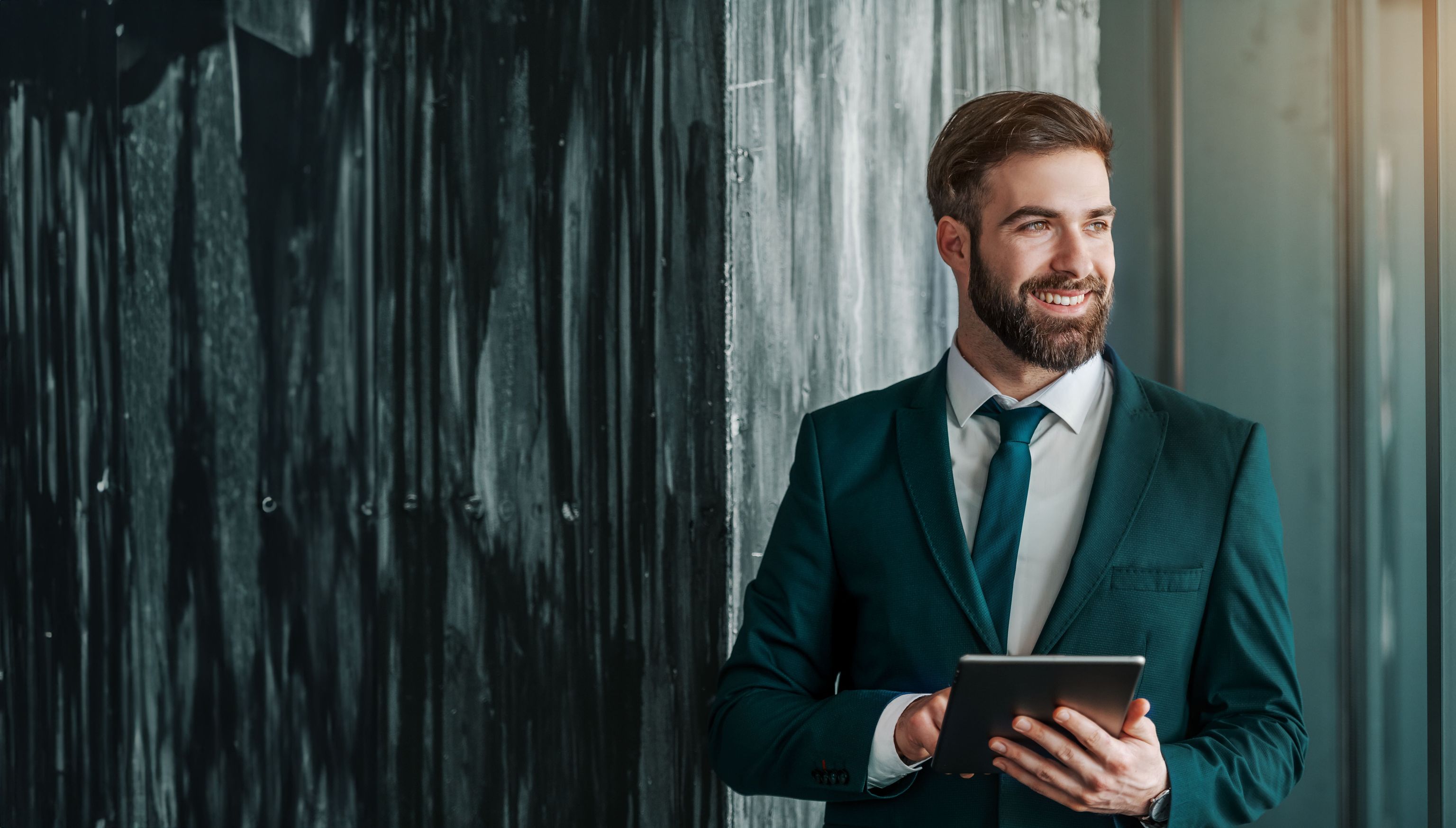 A smiling businessman in a dark green suit holding a tablet, standing in a stylish office with industrial and glass elements.