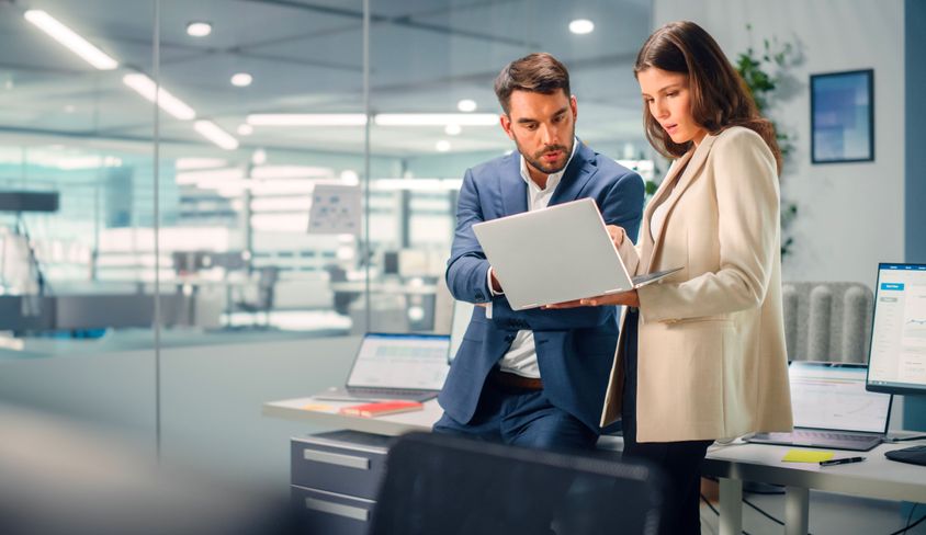 A male and female professional discussing data on a laptop in a high-tech office.